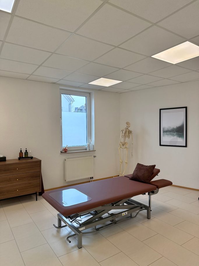 Behandlungsraum mit Liege, einem Fenster, einem Skelettmodell und einem Bild an der Wand.