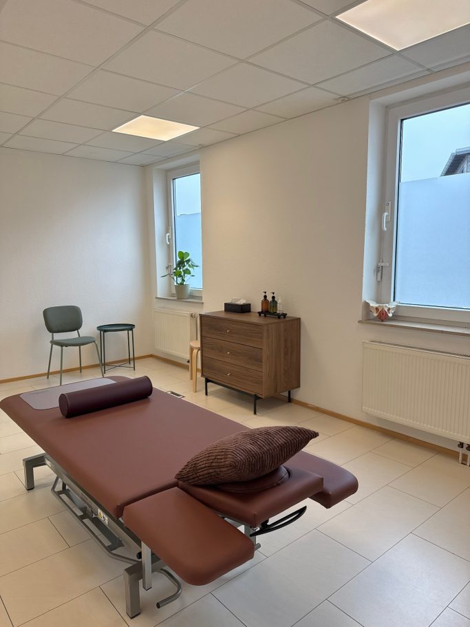 Behandlungsraum mit einer Liege, einem Stuhl und einem Sideboard, Pflanzen im Fenster.