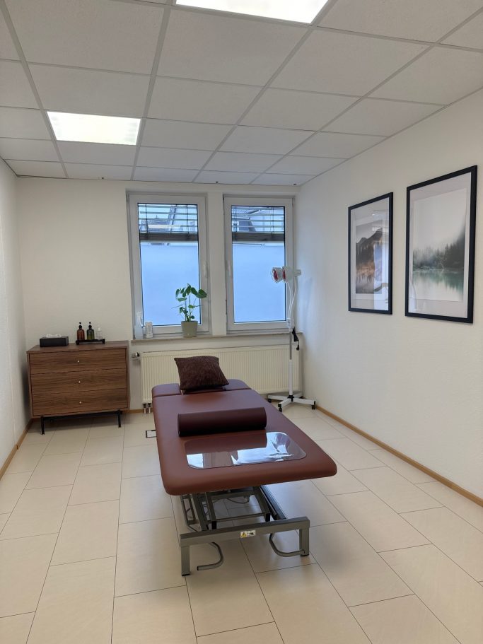 Behandlungsraum mit Liege, Fenstern und dekorativen Bildern an der Wand.